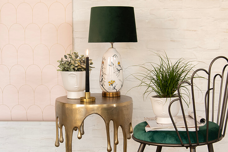 A modern interior with a gold-colored side table and on top sits a white flower pot, gold-colored candle holder, and a modern table lamp