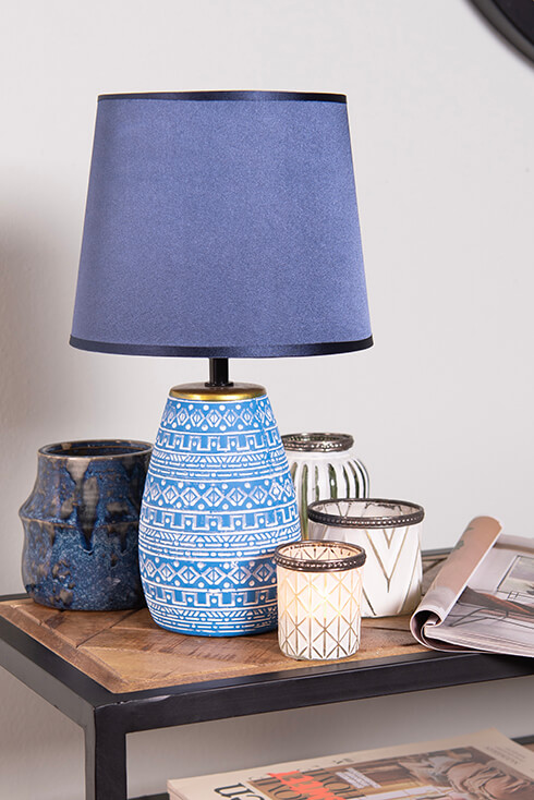 A blue table lamp with a blue lampshade surrounded by tea light holders and a flower pot