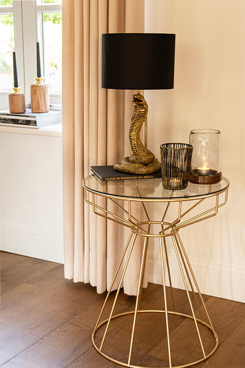 A gold-colored side table with a serpent table lamp with a black lampshade, a tea light holder, and glass lantern