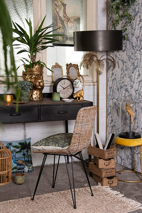 A botanical space with various home accessories, a wicker chair with throw pillows, and a floor lamp with gold-colored palm leaves and a black lampshade