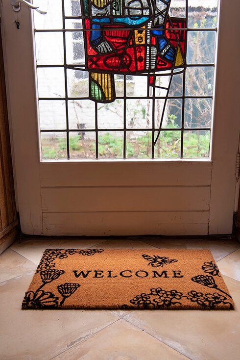 A doormat indoors with 'welcome' on it and various flowers and bees