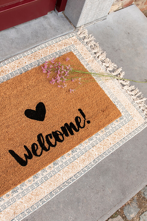 A doormat with 'welcome' and a heart on it, and underneath the doormat is a rectangular rug