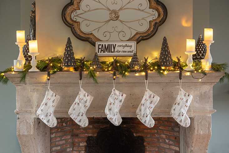 A large mantelpiece decorated with Christmas trees, Christmas lights, and deer Christmas stocking hangers with white Christmas stockings hanging from them