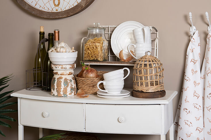 A white cabinet with a bottle rack, cups, plates, aprons, a basket, and a glass jar of pasta