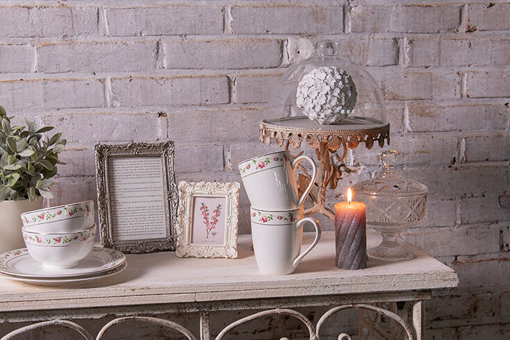 Une cheminée avec une boîte à bijoux, une cloche en verre, des tasses, des bols, des assiettes, des cadres photo, une bougie et un pot de fleurs