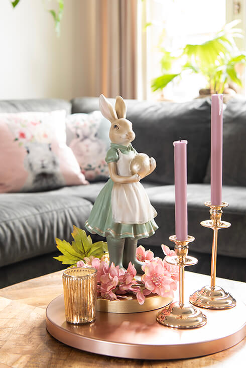 Een salontafel met een konijnen beeld, twee goudkleurige kandelaren met pastelpaarse kaarsen erin en roze kunstbloemen