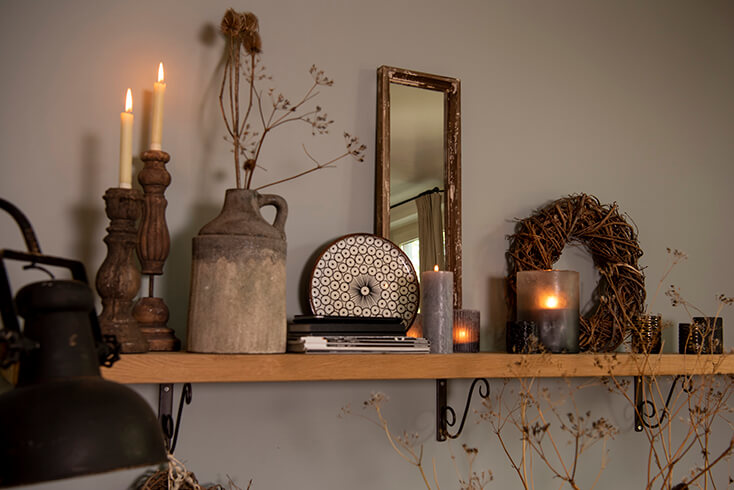 Ein rustikales Wandregal mit Regalträgern darunter und rustikalen Wohnaccessoires oben drauf, darunter ein Spiegel, eine Kanne, Kränze, Kerzenständer und Teelichthalter
