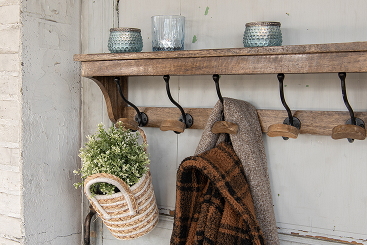 Eine rustikale Holzgarderobe, an der ein Korb mit einer Pflanze hängt, eine Jacke mit einem Schal und blaue Teelichthalter auf der Garderobe stehen