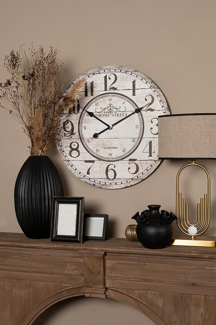 A mantelpiece with flower pots, photo frames, and a table lamp, and in the background, a large white wall clock