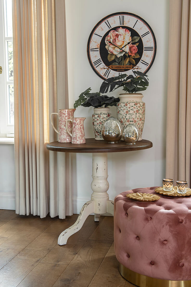 A pink pouf, a side table with flower pots and glass boxes, and in the background, a wall clock with a flower in the middle