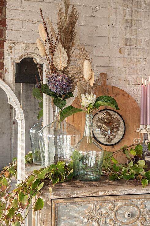 Een kastje met daarop kandelaars, spiegels, wanddecoratie en vazen met droogbloemen en kunstplanten