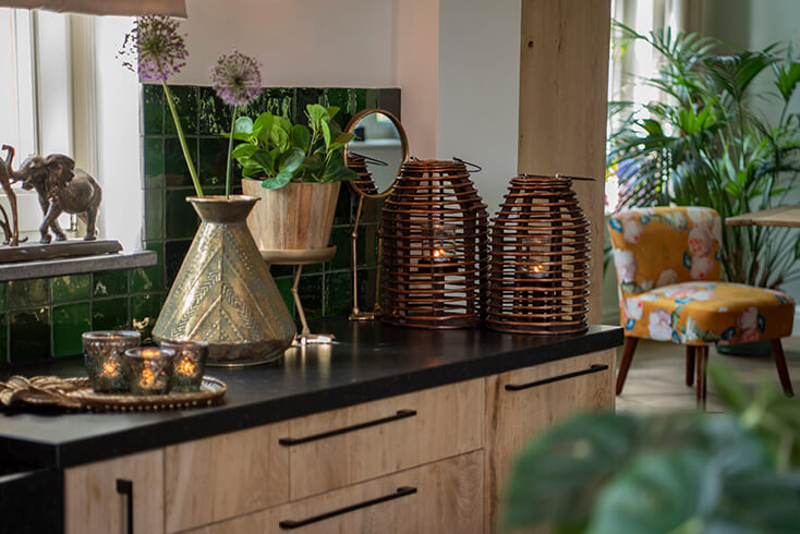 Un bancone con varie decorazioni per la casa, tra cui due lanterne botaniche, uno specchio da tavolo, un vaso in metallo e un vaso da fiori in legno