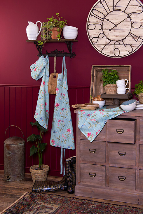 A rug, a cabinet with bowls, plates, and mugs. Also, hanging on a coat rack are aprons and oven mitts; on the wall hangs a clock, and to the left is a trash can