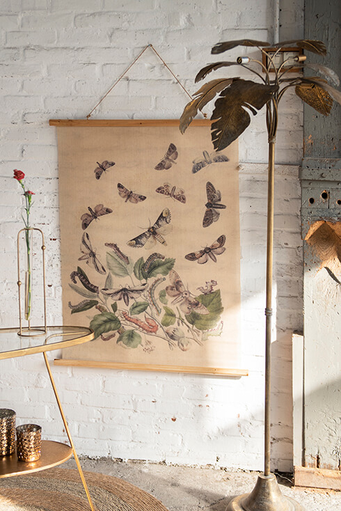 A side table and a lamp in the foreground, and in the background a wall tapestry of butterflies