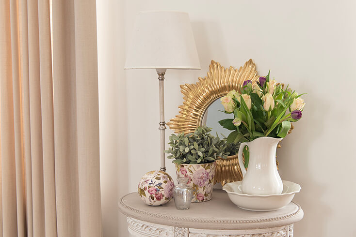 Een romantische bijzettafel met daarop een witte schenkkan, romantische bloempot, moderne wandspiegel en een landelijke tafellamp