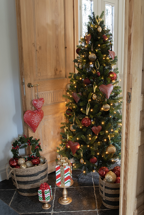 Een traditionele kerstboom met rode en goudkleurige kerstballen en kersthangers en ervoor staat een mand vol kerstballen