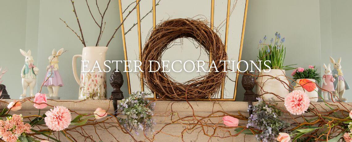 A serene and tasteful Easter decor adorns a fireplace mantel. At its center lies a natural wreath crafted from twigs, flanked by long, slender branches that add height and an organic feel to the arrangement. On one side of the wreath sits a large, white pitcher adorned with a floral pattern, contributing to the springtime atmosphere. Scattered throughout the scene are various Easter bunny figurines, each sporting their own unique outfit and pose, adding a playful and festive accent. Nestled amongst these decorations are pots of fresh spring flowers such as hyacinths and daffodils, their vibrant colors and lush foliage enhancing the seasonal charm. Completing the setup are scattered silk flowers and twigs seamlessly blending with the fresh flora. Above this enticing arrangement, the words "EASTER DECORATIONS" are displayed, signaling the theme of Easter adornments. The overall effect is that of a carefully curated collection evoking the freshness and renewal of spring.