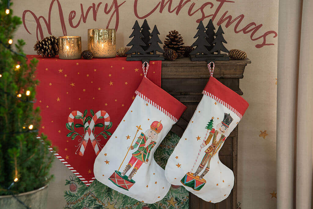A festive Christmas decoration, focusing on a mantelpiece where two Christmas stockings are hung. The stockings feature illustrations of nutcrackers in traditional costumes, with a border of red and white stripes along the top. The background is a red cloth with golden stars and the text "Merry Christmas" in ornate letters. On the mantelpiece are two golden candle holders casting a warm glow, next to a wooden decoration of pine trees and pine cones, emphasizing the natural theme of the season. The illuminated Christmas tree on the left side adds an extra cozy atmosphere to the scene.