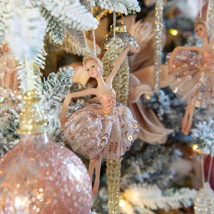 A Christmas tree ornament in the shape of a ballerina. The ballerina is dressed in a sparkling, pink tutu adorned with sequins and wears a matching tiara. Her arms are gracefully raised, and she has a delicate, joyful expression. In the background, you can see more glittering decorations and a second ballerina ornament. The Christmas tree itself is richly adorned with white and light pink tones, creating a soft and enchanting atmosphere. The focus is on the ballerina in the foreground, with a gentle blur in the background, giving a dreamy effect.