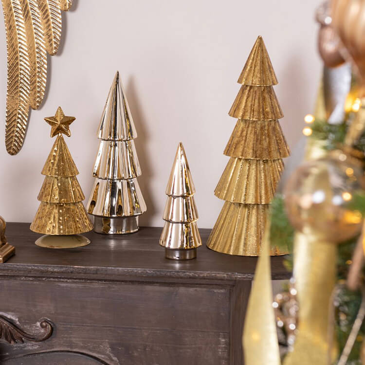 Een verzameling van decoratieve kerstbomen te zien op een donkere, houten console of dressoir. De bomen zijn gemaakt van verschillende materialen en in verschillende stijlen en formaten, sommige met een glanzende afwerking, andere met een getextureerd of gehamerd effect. De kleuren variëren van goud tot zilver, wat zorgt voor een modern en stijlvol kleurenpalet. Een deel van een grotere, gouden kerstdecoratie is zichtbaar aan de rechterkant van de foto, wat suggereert dat deze display deel uitmaakt van een grotere kerstsfeersetting. De algehele uitstraling is eigentijds en elegant, passend bij een verfijnde kerstdecoratie.