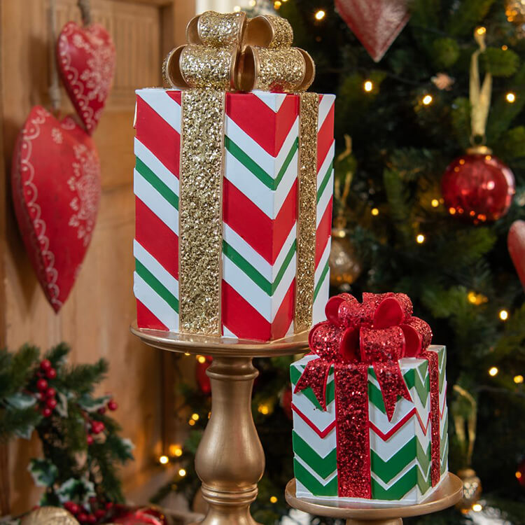 Twee decoratieve cadeau-achtige figuren geplaatst op een gouden standaard. De figuren zijn versierd met een traditioneel kerstthema: het grotere 'cadeau' bovenaan heeft rode en witte strepen met gouden glitterbanden en een gouden glitterstrik. Het kleinere 'cadeau' beneden heeft een vergelijkbaar ontwerp met een rode glitterstrik. Beide decoraties staan voor een achtergrond van een rijkelijk versierde kerstboom, compleet met lichtjes en rode kerstballen. Links in de afbeelding zie je nog een kerstdecoratie in de vorm van een rode kersthanger. Het geheel straalt een feestelijke en traditionele kerstsfeer uit.