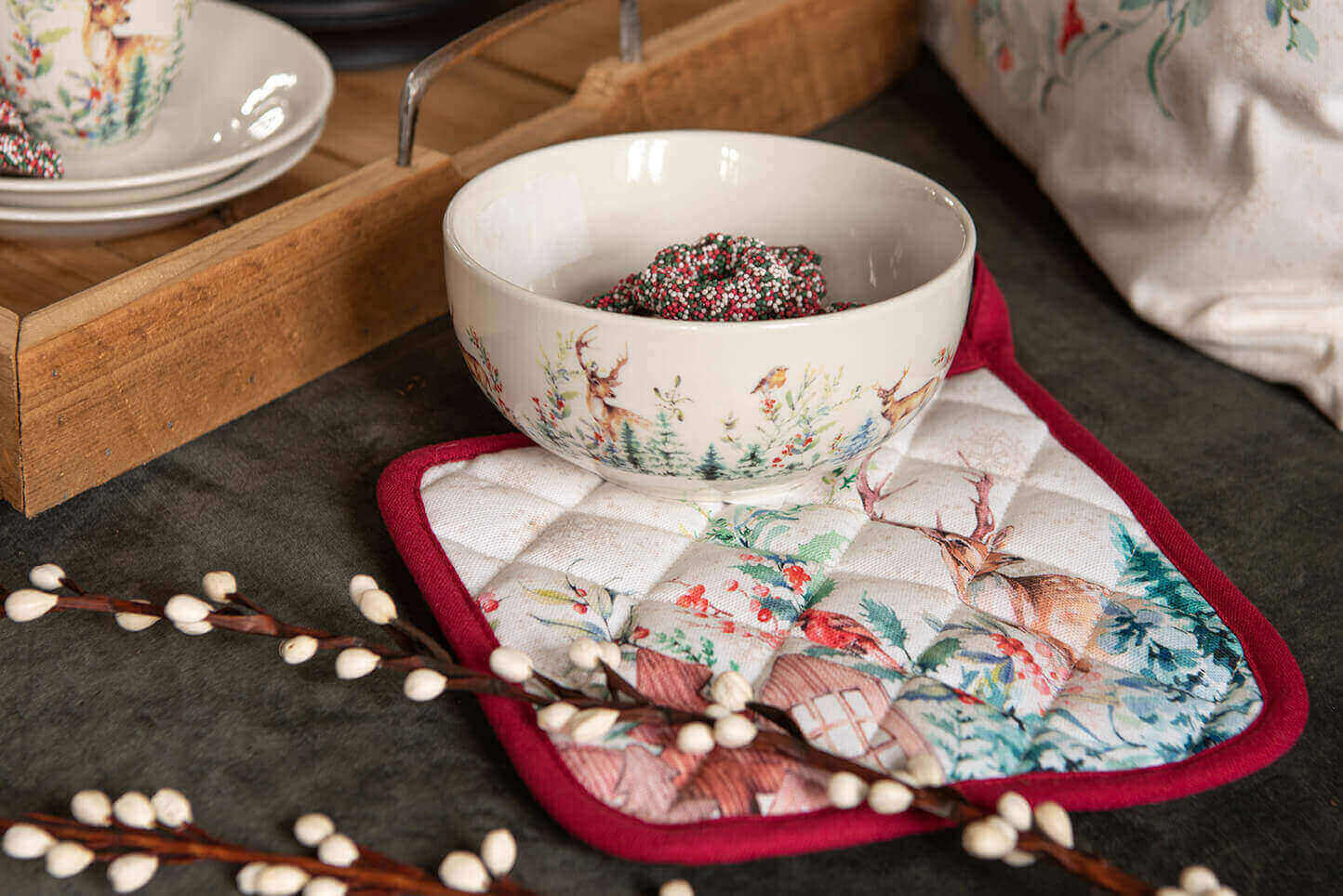 A festively set table with a focus on Christmas dinnerware. In the center sits a round bowl with a Christmas scene of deer and trees on the exterior, filled with what appears to be gingerbread or a similar festive treat. Underneath the bowl lies a pot holder with a similar rustic scene, bordered in red, adding to the Christmas atmosphere. In the background, we see more dinnerware with a matching design, likely part of a set. On the left side of the photo, there's a branch adorned with what appears to be cotton balls, giving a natural and wintry feel. In the top right corner of the photo, a portion of a fabric item with a Christmas pattern is visible. The overall ambiance exudes a cozy and homely Christmas atmosphere.