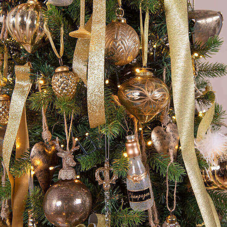 A close-up of a lavishly decorated Christmas tree. The tree is adorned with ornaments in a color scheme of gold and bronze, giving it a rich, luxurious appearance. Various types of decorations are visible, such as shiny and matte Christmas balls, sparkling garlands, stars, and unique ornaments like a heart, a reindeer, and even a champagne bottle. The details on the ornaments are refined, with textures and embellishments that reflect the light from the Christmas tree lights. All of this contributes to a festive and cheerful atmosphere typical of the Christmas season.