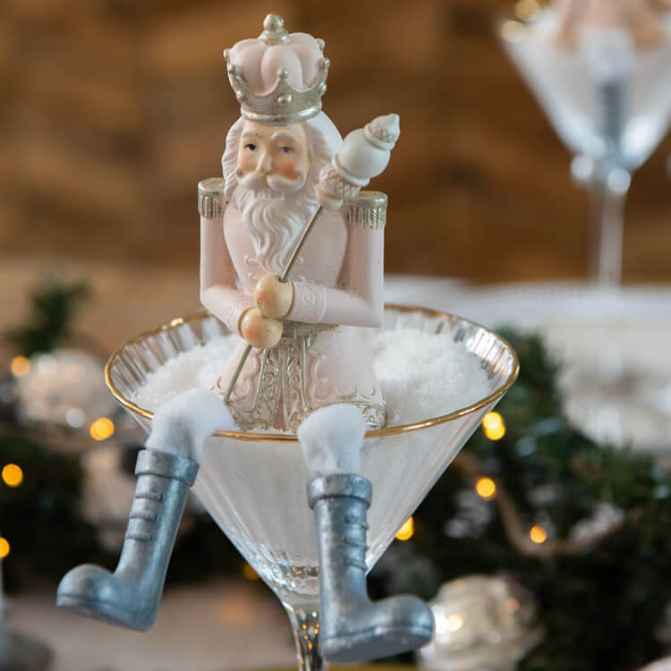 A graceful nutcracker figure, rendered in soft pastel colors, primarily pink and white, with gold and blue accents. The nutcracker wears a crown and holds a scepter. It stands in a martini glass filled with white decorative 'snow'. In the background, the faint outlines of a festively decorated space with lights and other Christmas decorations are visible. The scene exudes a luxurious and festive Christmas atmosphere.