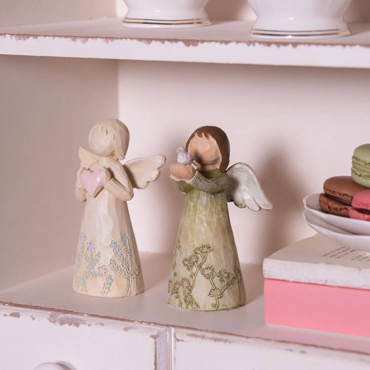 In the photo, you see two angel figurines placed on a light pink shelf, part of a white, shabby chic cabinet or rack. The angels have a rustic appearance, with soft pastel hues and delicate patterns on their robes. The angel on the left holds a heart, while the angel on the right appears to cradle a bird in her hands. On the right side of the shelf, there is a white tray with colored macarons, and next to the angels are a stacked pink box and a book, adding to the soft and serene decor. The overall atmosphere of the photo is gentle and tranquil.