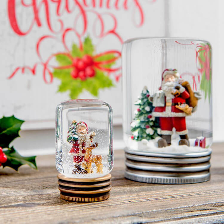 The image depicts two snow globes on a wooden surface. The front snow globe is smaller and shows a figure of Santa Claus with a reindeer, surrounded by falling 'snow'. This globe sits on a brown base with gold accents. The larger snow globe behind it depicts a scene with Santa Claus, a Christmas tree, and a sack of gifts. This globe has a wider silver-colored base. Behind the snow globes, a piece of decor with Christmas ornaments is visible, including holly leaves and the text "Merry Christmas" in red decorative letters, as well as a traditional Christmas rose illustration. The overall composition creates a cozy Christmas scene.