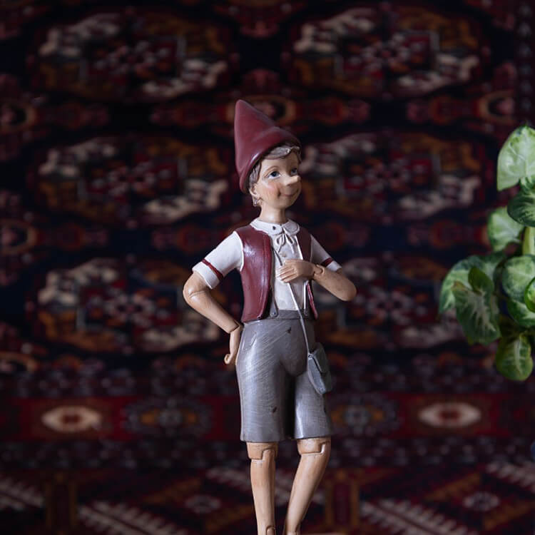 The photo depicts a character reminiscent of a fairytale figure, possibly an elf or a character from a classic story. The figure wears a traditionally styled outfit with a pointed hat, a white blouse with a front lace-up, a reddish-brown vest, and gray shorts. It stands with hands on hips and has a cheerful expression on its face. In the background, a colorful, detailed carpet is visible, adding to the atmosphere of a story or an old-fashioned setting. There is a vague green element on the right side of the image resembling a plant leaf, suggesting that this character may be situated in an indoor scene. The character appears carefully crafted and detailed, adding character and charm to the composition.