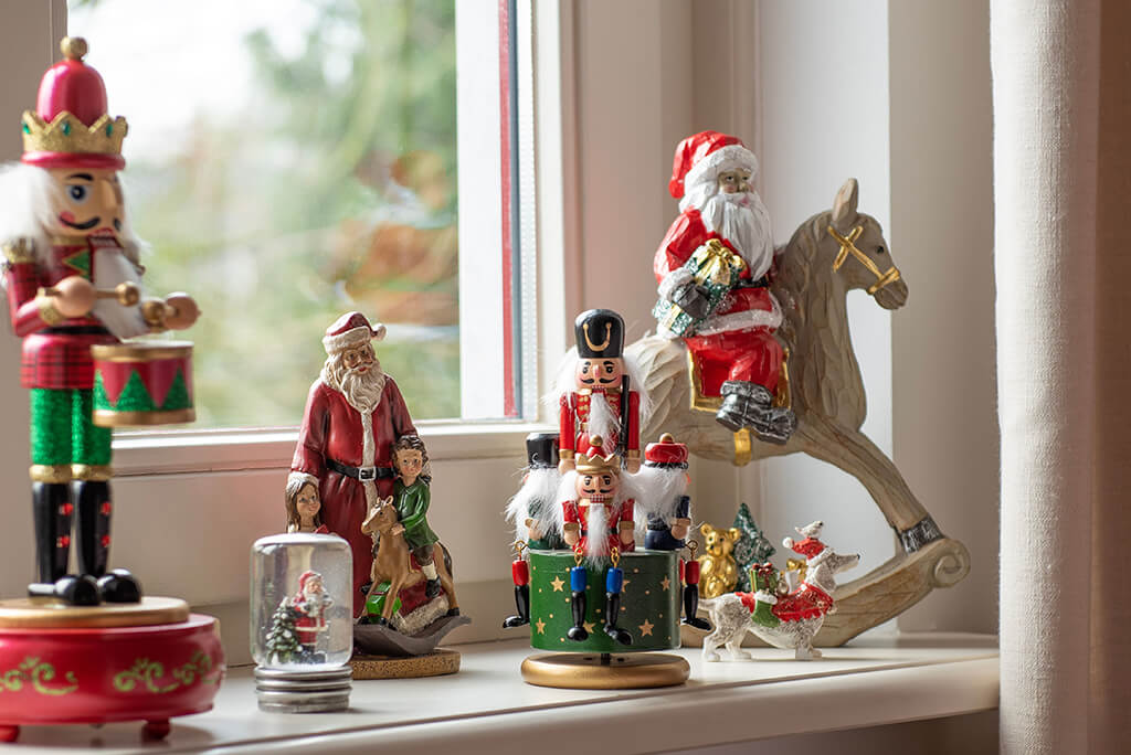 In the photo, there is a collection of Christmas decorations on a windowsill, with daylight streaming in through the window. On the left stands a large nutcracker figure wearing a red and green uniform and a crown, holding onto a pair of drums. Next to him is a Christmas figure, likely Santa Claus, in a red costume, along with child figures and a smaller snow globe with a Christmas scene inside. In the middle is a stackable nutcracker on a green box with a drum, and a smaller Santa Claus figure beside it. On the right side is a decorative piece featuring Santa Claus on a rocking horse. Smaller ornaments, including a mini Christmas tree and a reindeer figure, complete the scene. This collection creates a traditional and cozy Christmas atmosphere.