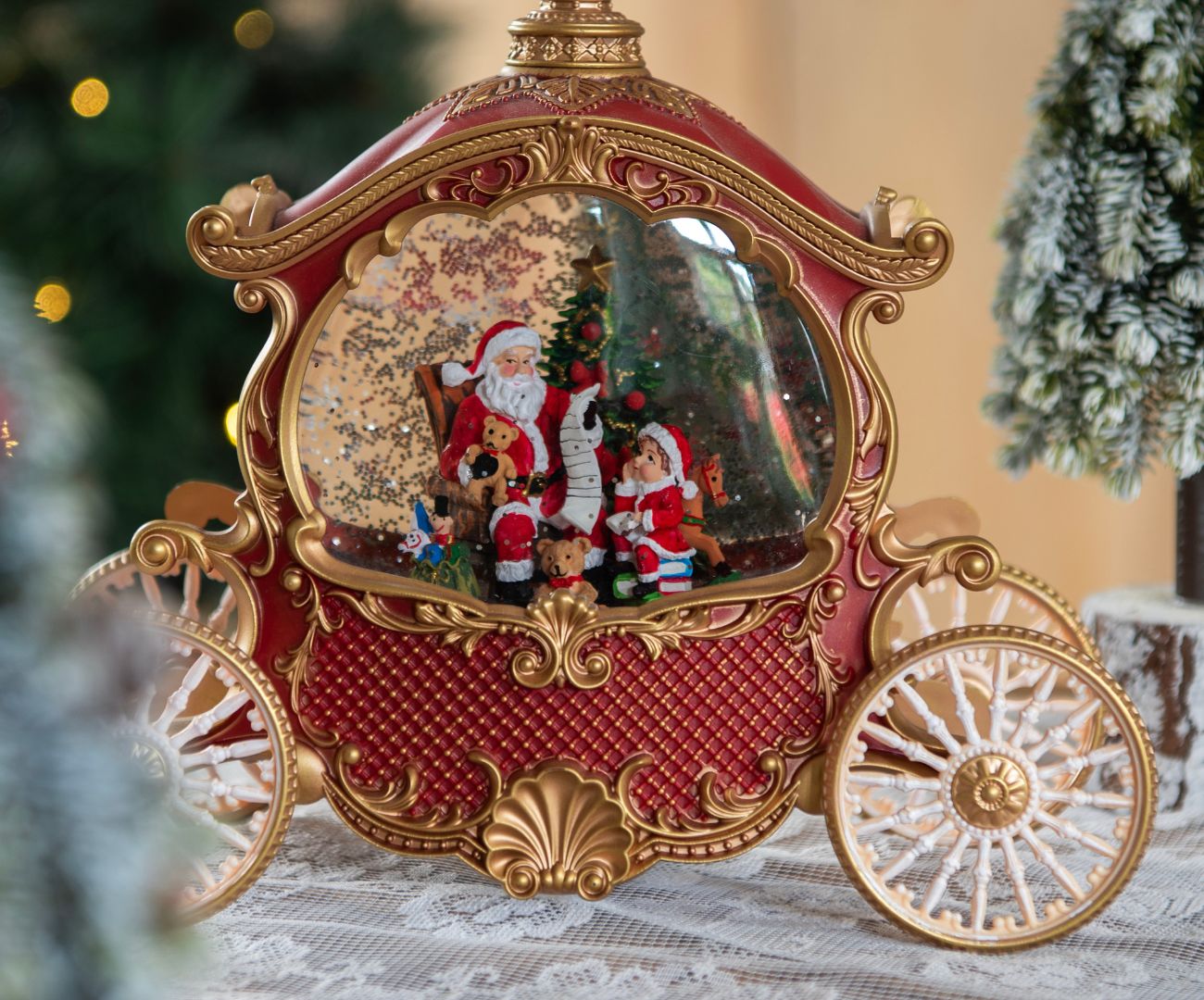 The image depicts two snow globes on a wooden surface. The front snow globe is smaller and shows a figure of Santa Claus with a reindeer, surrounded by falling 'snow'. This globe sits on a brown base with gold accents. The larger snow globe behind it depicts a scene with Santa Claus, a Christmas tree, and a sack of gifts. This globe has a wider silver-colored base. Behind the snow globes, a piece of decor with Christmas ornaments is visible, including holly leaves and the text "Merry Christmas" in red decorative letters, as well as a traditional Christmas rose illustration. The overall composition creates a cozy Christmas scene.