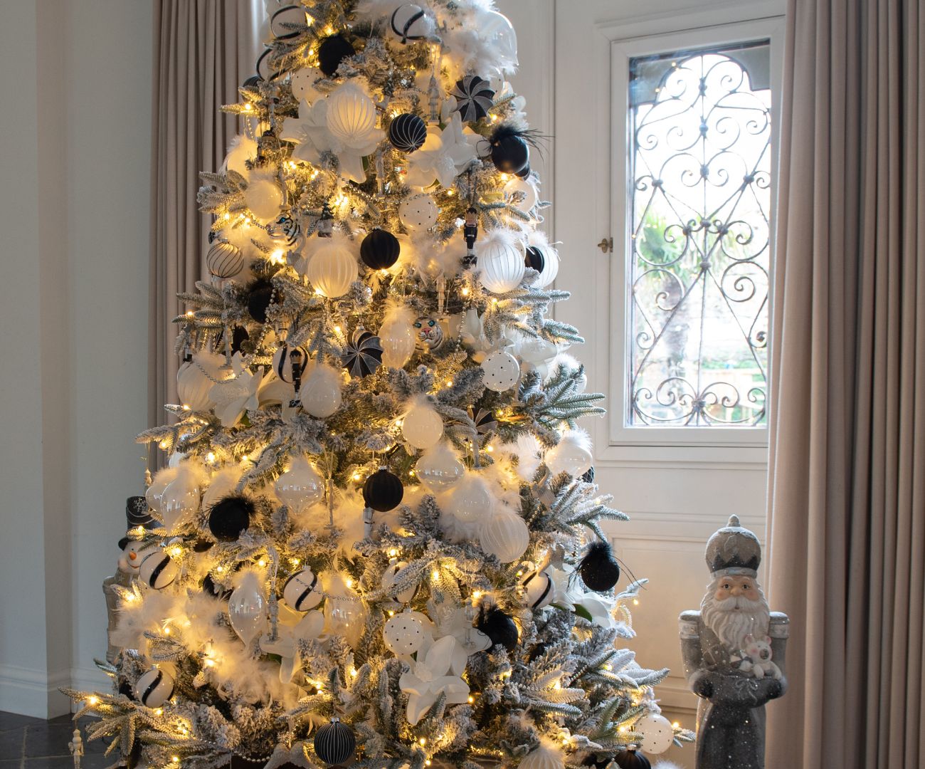 Two decorative gift-like figures placed on a golden stand. The figures are adorned with a traditional Christmas theme: the larger 'gift' at the top features red and white stripes with golden glitter ribbons and a golden glitter bow. The smaller 'gift' below has a similar design with a red glitter bow. Both decorations stand against a background of a lavishly decorated Christmas tree, complete with lights and red baubles. On the left side of the image, another Christmas decoration in the form of a red Christmas ornament is visible. The whole scene exudes a festive and traditional Christmas atmosphere.