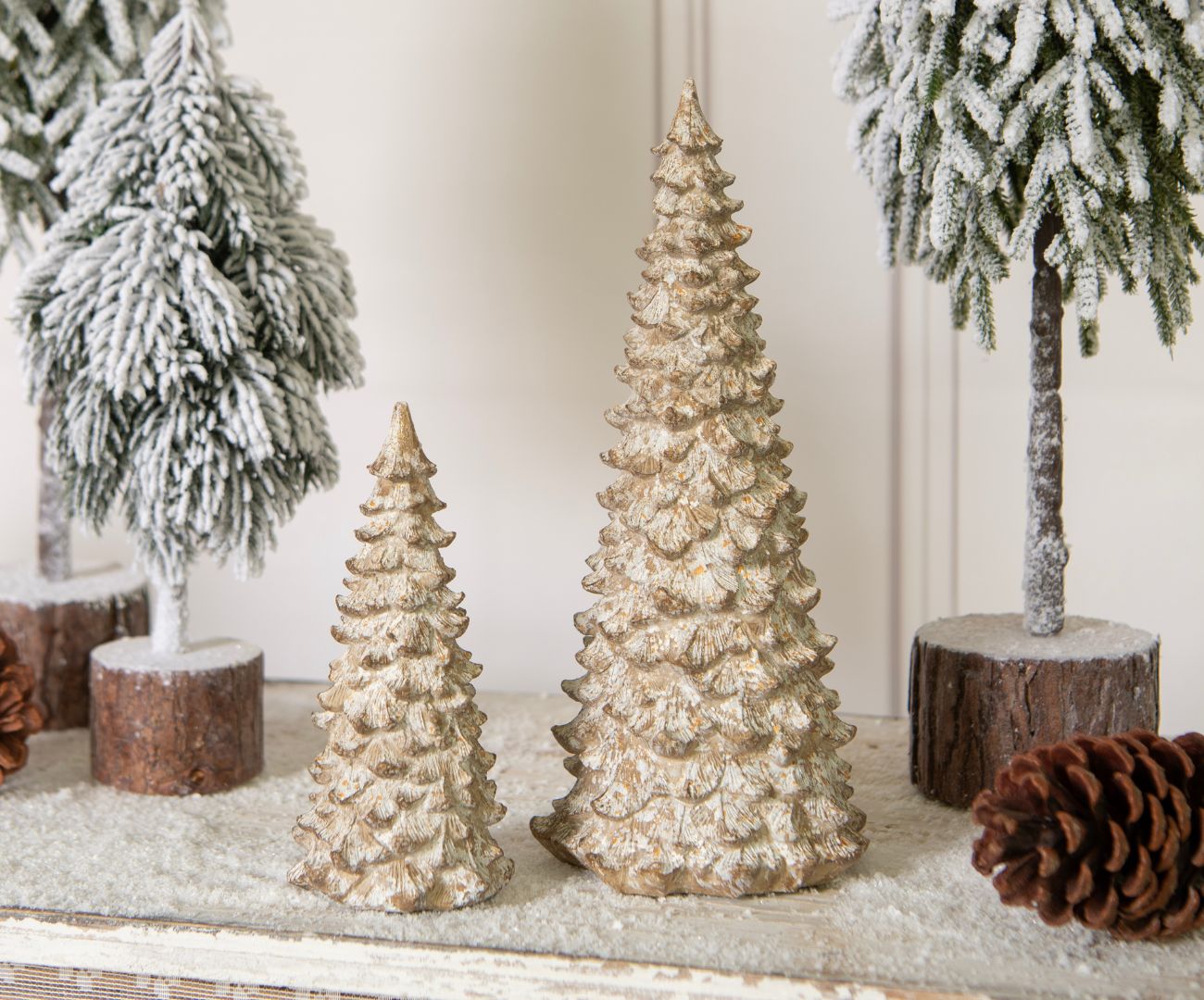 A collection of decorative Christmas trees is displayed on a dark wooden console or sideboard. The trees are made of various materials and in different styles and sizes, some with a glossy finish, others with a textured or hammered effect. The colors range from gold to silver, creating a modern and stylish color palette. Part of a larger, golden Christmas decoration is visible on the right side of the photo, suggesting that this display is part of a larger Christmas decor setting. The overall look is contemporary and elegant, fitting for sophisticated Christmas decor.