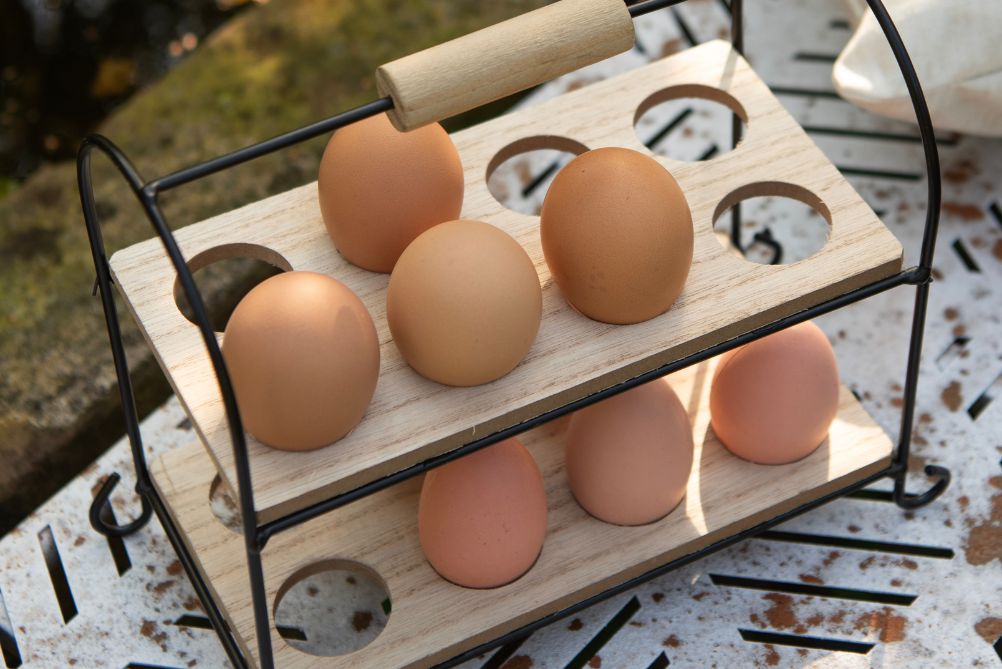 Eine stimmungsvolle Osterfrühstücksszene. Im Mittelpunkt steht ein leuchtend rosa bemaltes Osterei in einem weißen Eierbecher mit einem Hasenmotiv. Neben dem Eierbecher liegen ein Teller und eine Tasse, beide verziert mit subtilen Hasenillustrationen und Blattmotiven, die eine weiche und festliche Atmosphäre schaffen. Auf der linken Seite des Fotos ist ein Teil eines köstlich aussehenden Croissants zu sehen, was der Szenerie eine einladende Note verleiht. Die Gegenstände sind auf einem Tischtuch mit einem groß abgebildeten Hasen platziert, was die Kohärenz des Osterthemas unterstreicht. Im Hintergrund sind weitere Ostereier und ein Teil eines Kranzes zu sehen, was die Frühlingsstimmung weiter verstärkt. Die gesamte Anordnung strahlt Wärme und Gemütlichkeit aus, perfekt für ein festliches Osterfrühstück.