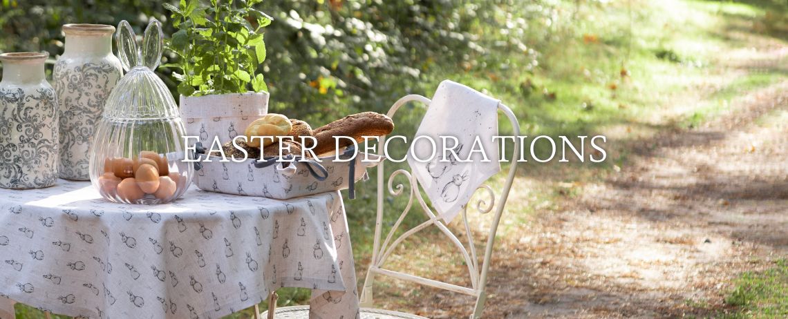 A serene and tasteful Easter decor adorns a fireplace mantel. At its center lies a natural wreath crafted from twigs, flanked by long, slender branches that add height and an organic feel to the arrangement. On one side of the wreath sits a large, white pitcher adorned with a floral pattern, contributing to the springtime atmosphere. Scattered throughout the scene are various Easter bunny figurines, each sporting their own unique outfit and pose, adding a playful and festive accent. Nestled amongst these decorations are pots of fresh spring flowers such as hyacinths and daffodils, their vibrant colors and lush foliage enhancing the seasonal charm. Completing the setup are scattered silk flowers and twigs seamlessly blending with the fresh flora. Above this enticing arrangement, the words "EASTER DECORATIONS" are displayed, signaling the theme of Easter adornments. The overall effect is that of a carefully curated collection evoking the freshness and renewal of spring.