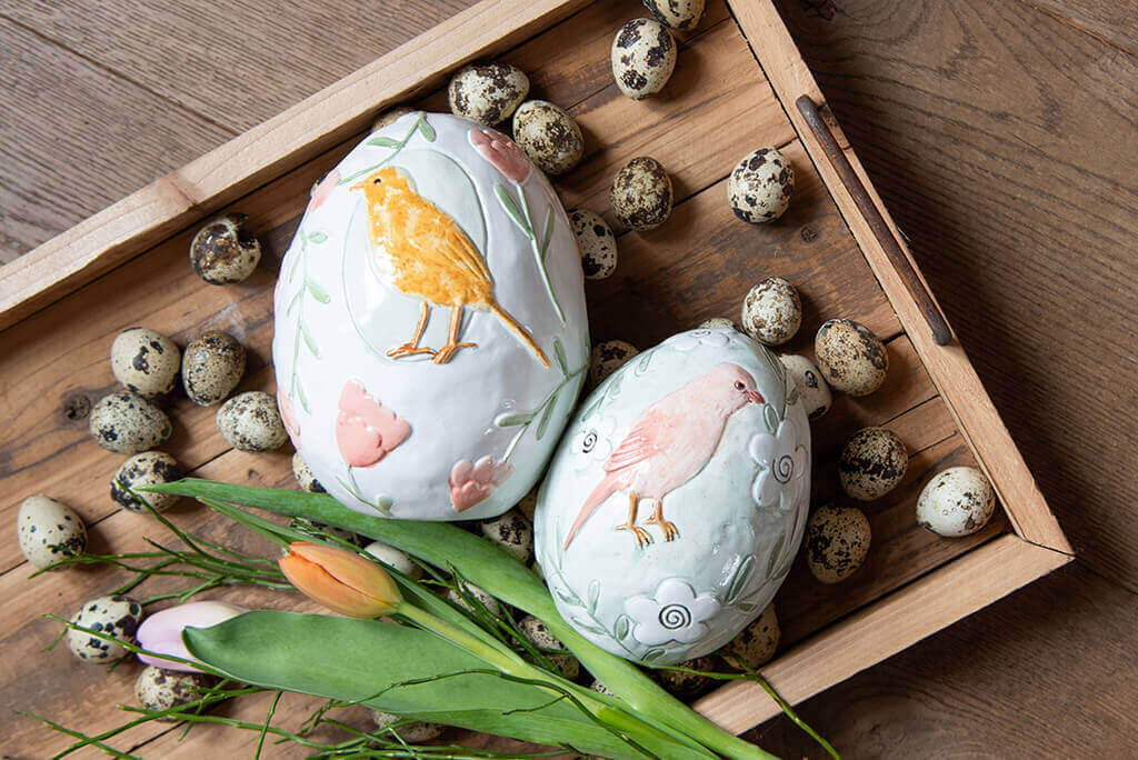 An artistic Easter decoration arranged on a wooden surface. At the center are two large decorative eggs, painted with lovely images of an orange bird and a pink bird, surrounded by subtle green leaves and blossom motifs. A fresh orange tulip lies between the eggs. Multiple quail eggs are casually scattered around the eggs, giving a natural and rustic appearance. The entire composition is placed in a wooden tray with raised edges, adding depth and texture to the image. The overall effect evokes a sense of spring and Easter joy.