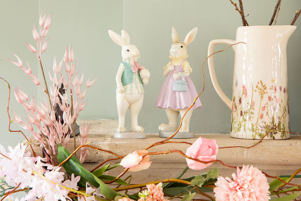 The picture depicts a charming Easter decoration consisting of two graceful porcelain bunny figures, adorned in pastel-colored attire - one in a light green vest and bowtie, and the other in a lovely pink dress with a matching purse. Beside them stands a large, cream-colored pitcher with a delicate floral motif symbolizing spring. The bunnies and the pitcher are placed on a rustic wooden surface that complements the natural theme. The scene is enriched with various spring flowers and branches, both subtle and colorful, including pink buds and delicate pink blossoms scattered around, creating a soft and festive atmosphere. The overall ambiance exudes a sense of serenity and the celebration of spring.