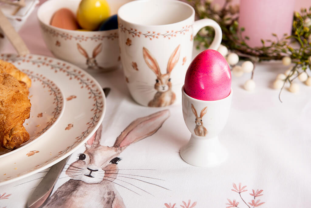 A charming Easter breakfast scene is depicted. At the center sits a bright pink painted Easter egg in a white egg cup with a rabbit motif. Adjacent to the egg cup are a plate and a mug, both adorned with subtle rabbit illustrations and leaf motifs, creating a soft and festive ambiance. On the left side of the photo, a portion of a delicious-looking croissant is visible, adding an inviting touch to the setting. These items are placed on a tablecloth featuring a large depicted rabbit, emphasizing the cohesion of the Easter theme. In the background, more Easter eggs and a portion of a wreath are visible, further enhancing the springtime mood. The entire arrangement exudes warmth and coziness, perfect for a festive Easter breakfast.