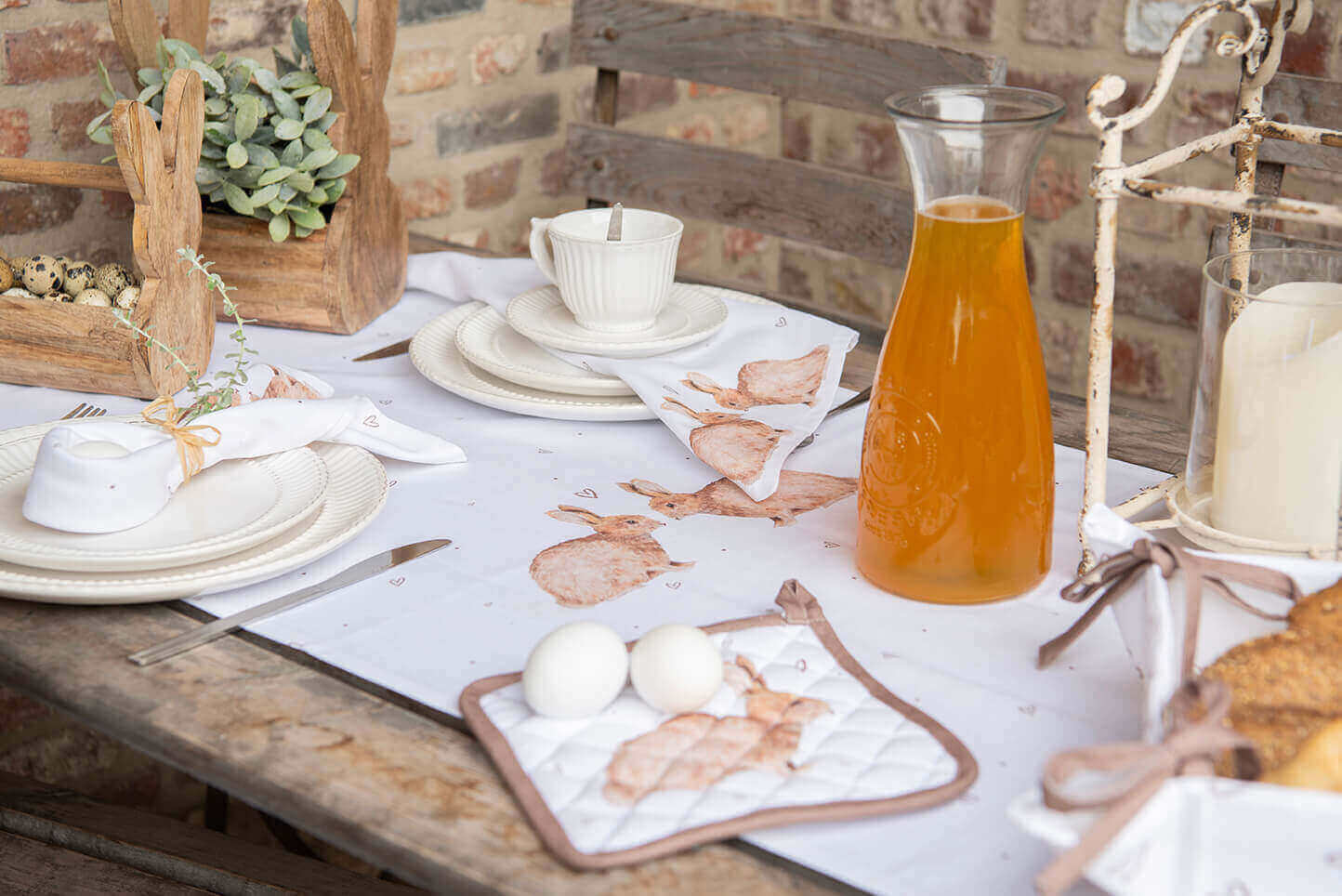 A rustic table setting with a countryside feel. On the table lies a white tablecloth adorned with playful rabbit illustrations. Cream-colored dinnerware is visible, featuring a subtle ribbed pattern along the edges. The napkins are neatly rolled and tied with a graceful string, placed on the plates. A coffee cup is stacked atop a matching saucer, evoking the inviting atmosphere of a coffee break.

Adjacent to the dinnerware sits a clear pitcher filled with orange liquid, radiating a refreshing vibe. A decorative, weathered metal candle holder with a large candle inside contributes to the cozy ambiance. On the left side of the table stands a wooden centerpiece adorned with inlaid quail eggs beside a pot of vibrant green plants.

The scene exudes a simple yet sophisticated atmosphere, perfect for a relaxed Easter breakfast or brunch outdoors. The rustic wooden furniture and brick wall in the background add to the charming, countryside appeal of the photo.