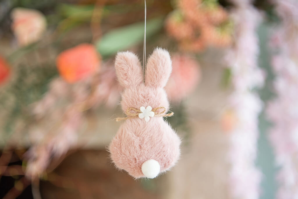 A cute, plush Easter bunny ornament in soft pink, suspended in the air. The bunny is shown from behind, focusing on its large, fluffy ears and a small tail peeking out at the bottom. Around the bunny's neck is a rustic jute string tied with a small, white floral decoration in the middle, adding a touch of rural charm. The background is soft and blurry with hints of green and pink, suggesting the bunny hangs against a backdrop of spring flowers. This scene exudes a sense of Easter and the joyfulness of the season.