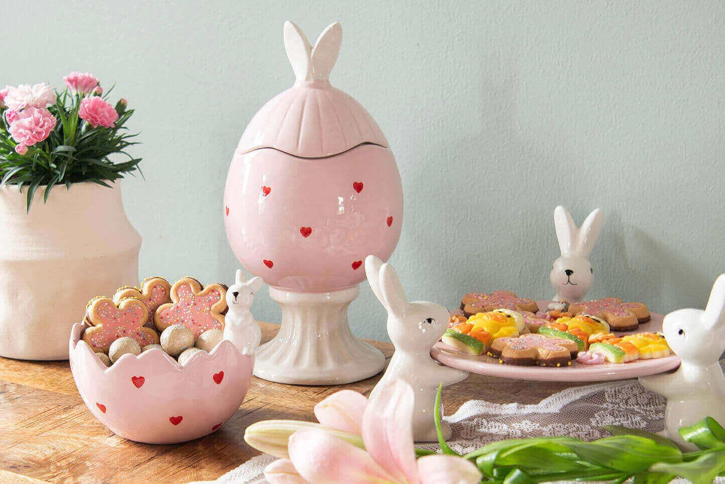 The photo depicts a charming Easter table setting. In the foreground is an adorable, pink flower-shaped bowl filled with heart-shaped cookies and small Easter eggs, with a small bunny figurine protruding from it. Next to the bowl sits a larger, elegant pink egg-shaped pot with bunny ears on the lid and adorned with small hearts. Beside it stands a vase filled with lush pink flowers, adding freshness and color to the scene. On the right side of the photo, you can see a pink plate filled with colorful canapés, surrounded by porcelain bunnies presenting the plate. The entire setting is placed on a wooden table and is enlivened by some spring flowers in the foreground, emphasizing the spring and Easter mood. The pastel colors and bunny theme offer a delightful and festive atmosphere characteristic of Easter.