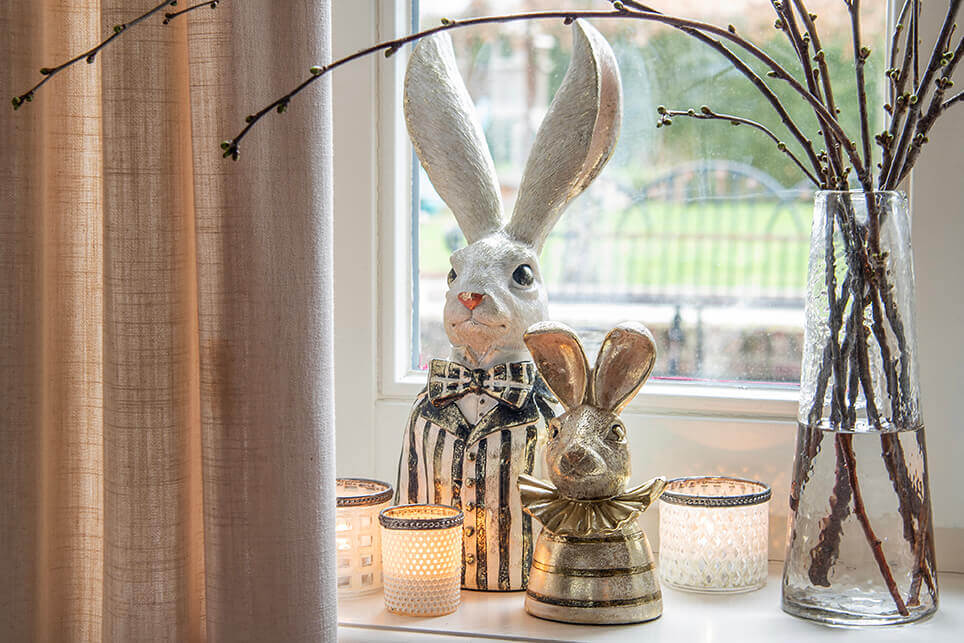 In front of the window stands a decorative arrangement featuring two rabbit figurines: one large, wearing a striped outfit and sporting striking, long ears, and one smaller, golden rabbit with a shiny bow. Between the rabbits are several ambiance lights emitting a warm glow, casting a cozy pattern on the surroundings. On the right side of the composition sits a glass vase filled with straight branches, adding a natural and rustic element to the scene. The ensemble creates a homely and festive atmosphere, fitting for the Easter season.
